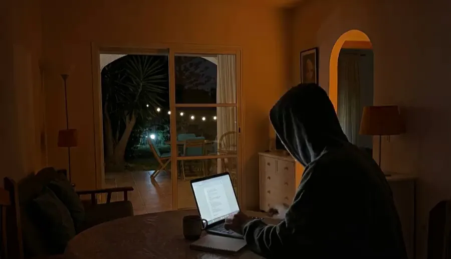Adam typing on a laptop at a round table in his living room at night, with a lit garden behind the glass door